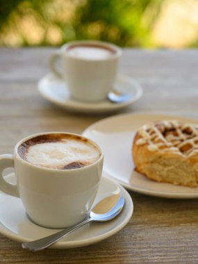 Fotoğrafta iki fincan kahveli hamur işleri var. Kahvaltı ahşap bir masanın üzerindeki bir kafede yer alır. Arka planda tropikal bitki örtüsü görünür..