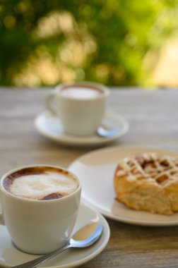 Fotoğrafta iki fincan kahveli hamur işleri var. Kahvaltı ahşap bir masanın üzerindeki bir kafede yer alır. Arka planda tropikal bitki örtüsü görünür..