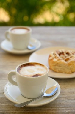 Fotoğrafta iki fincan kahveli hamur işleri var. Kahvaltı ahşap bir masanın üzerindeki bir kafede yer alır. Arka planda tropikal bitki örtüsü görünür..
