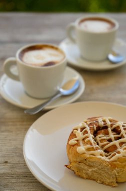 Fotoğrafta iki fincan kahveli hamur işleri var. Kahvaltı ahşap bir masanın üzerindeki bir kafede yer alır. Arka planda tropikal bitki örtüsü görünür..