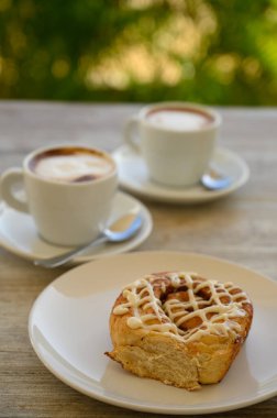 Fotoğrafta iki fincan kahveli hamur işleri var. Kahvaltı ahşap bir masanın üzerindeki bir kafede yer alır. Arka planda tropikal bitki örtüsü görünür..