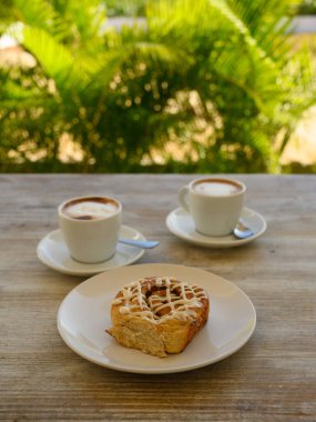 Fotoğrafta iki fincan kahveli hamur işleri var. Kahvaltı ahşap bir masanın üzerindeki bir kafede yer alır. Arka planda tropikal bitki örtüsü görünür..