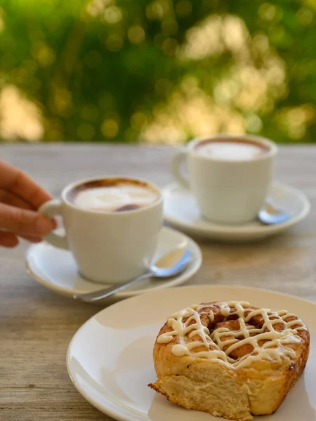 Fotoğrafta iki fincan kahveli hamur işleri var. Kahvaltı ahşap bir masanın üzerindeki bir kafede yer alır. Arka planda tropikal bitki örtüsü görünür..