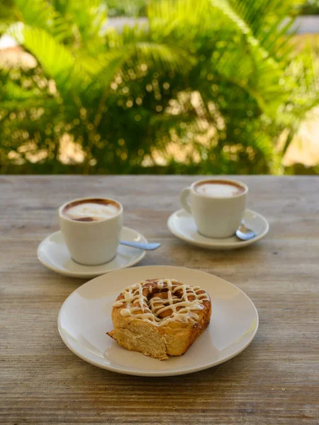 Fotoğrafta iki fincan kahveli hamur işleri var. Kahvaltı ahşap bir masanın üzerindeki bir kafede yer alır. Arka planda tropikal bitki örtüsü görünür..
