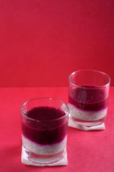 Photo shows vegan dessert in glass. Healthy food has two layers of white with chia seeds made from yogurt and top - pink, made from pitahaya juice. Pink color background.