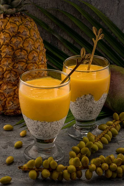 Photo shows a vegan dessert in glass. Healthy food has two layers: the bottom one is white with chia seeds made from yogurt and the top one is yellow, made from mango juice. In background is pineappe.