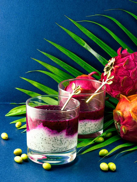 Photo shows vegan dessert in glass. Healthy food has two layers of white with chia seeds made from yogurt and top - pink, made from pitahaya juice. Several exotic fruits in background.