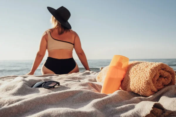 Концепція відпустки, сонцезахисний лосьйон та рушник у фокусі, вид ззаду жінки, що сидить на пляжі біля моря — стокове фото