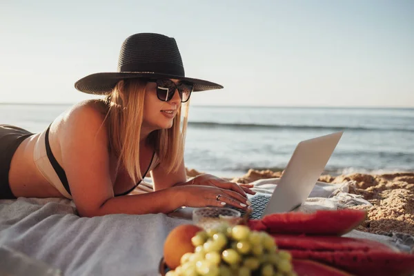 Mulher bonita em óculos de sol e chapéu usando laptop, desfrutando de piquenique em férias enquanto deitado em Sandy Beach by the Sea — Fotografia de Stock