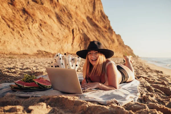 Jovem alegre em maiô e chapéu sorrindo para a câmera enquanto trabalhava no laptop em férias pelo mar — Fotografia de Stock