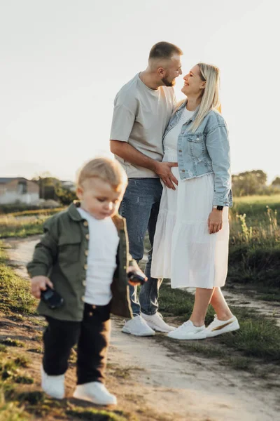 Щаслива молода сім'я на відкритому повітрі, Отець тримає свою вагітну дружину животом, поки їх Тоддлер син грає навколо них — стокове фото