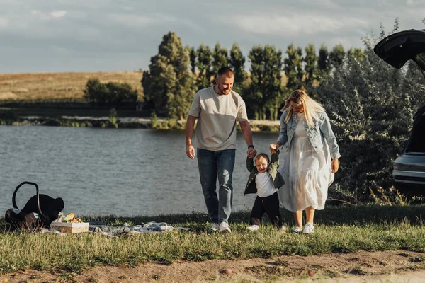 Madre y padre ayudan a su hijo pequeño a dar los primeros pasos, picnic familiar al aire libre junto al lago — Foto de Stock