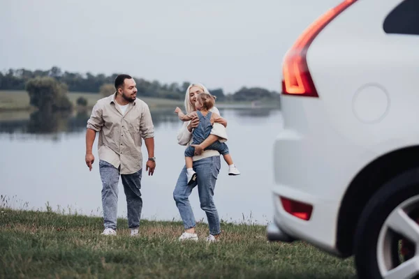 Feliz familia joven que se divierte al aire libre cerca del lago, mamá y papá con su hija pequeña están disfrutando de viaje por carretera en coche —  Fotos de Stock