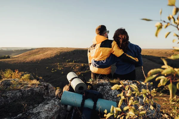 Blick zurück auf das Wanderpaar, das sich bei Sonnenuntergang umarmt und am Hügelrand sitzt, Reisepaar genießt seine Wanderung auf dem Gipfel des Hügels — Stockfoto