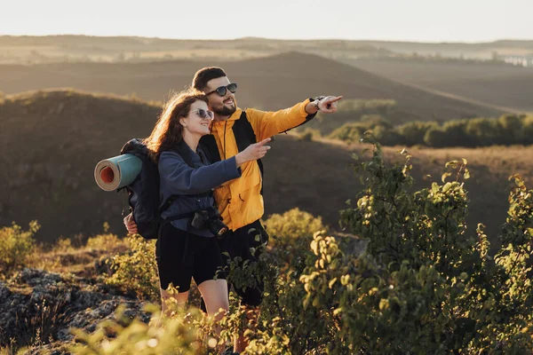 Glückliche Reisende: Mann und Frau umarmen sich bei Sonnenuntergang auf dem Hügel und zeigen etwas, das sie überrascht hat — Stockfoto
