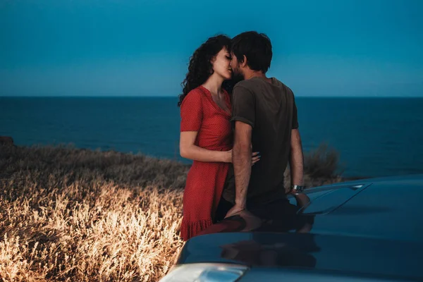 Unga par man och kvinna kyssas och kramas nära sin bil på kustlinjen vid Nightfall — Stockfoto