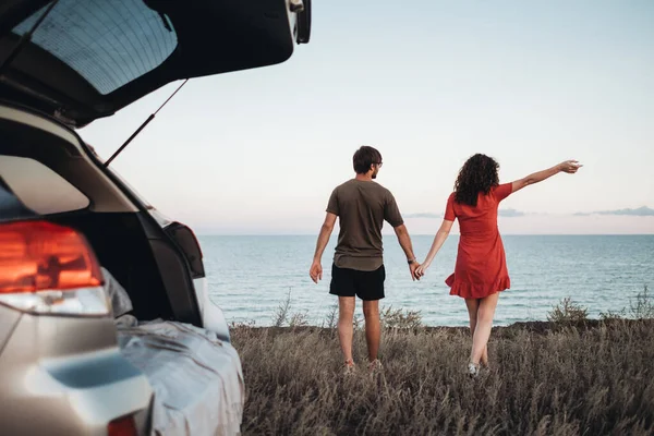 在大海的背景下手挽手散步的年轻夫妇，享受乘车旅行的乐趣 — 图库照片