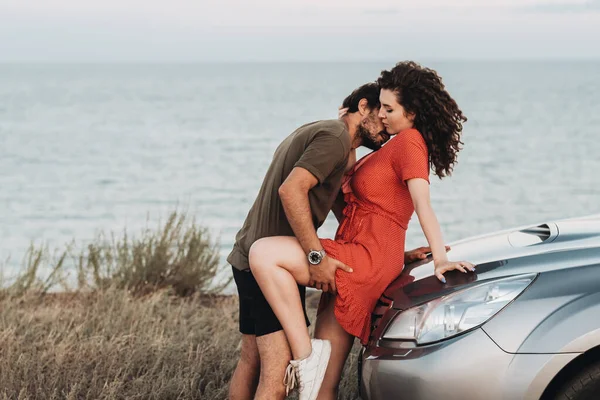 Νεαρό ζευγάρι στο οδικό ταξίδι, ο άνθρωπος Intimate Φιλί γυναίκα στο λαιμό για την κουκούλα του αυτοκινήτου SUV τους στο φόντο της θάλασσας στο ηλιοβασίλεμα — Φωτογραφία Αρχείου