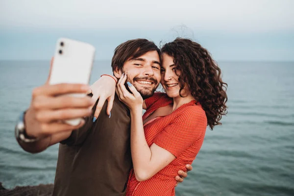 Happy kaukaski para mężczyzna i kręcone Brunetka kobieta przytula się stojąc przed morzem i robi selfie na smartfon — Zdjęcie stockowe