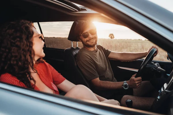 Hombre alegre y mujer morena rizada sentados en el coche, pareja joven disfrutando de un viaje por carretera al atardecer —  Fotos de Stock