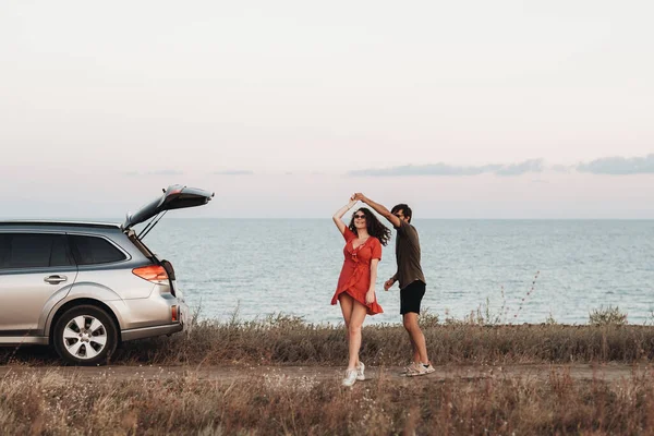 Pareja de jóvenes divirtiéndose en el fondo del mar, el hombre y la mujer bailando cerca de su coche SUV al atardecer —  Fotos de Stock