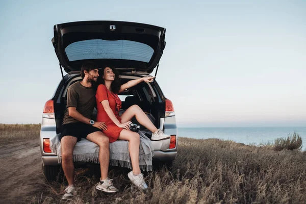 Man and Woman Sitting Inside Opened Trunk of Their SUV Car, Young Couple Enjoying Road Trip Along Sea at Sunset — Stok Foto