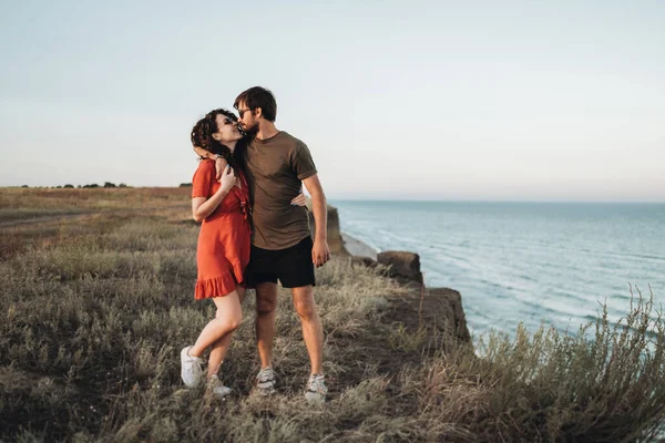 Fröhliches junges Paar steht auf dem Hintergrund des Meeres, glücklicher Mann umarmt lockige Frau im roten Kleid bei Sonnenuntergang — Stockfoto