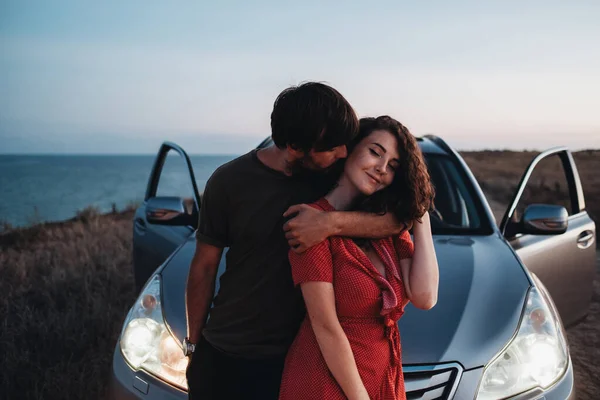 Joven pareja hombre y mujer abrazando y besándose cerca de su coche SUV en la playa después de la puesta del sol, amantes disfrutando de momentos juntos — Foto de Stock