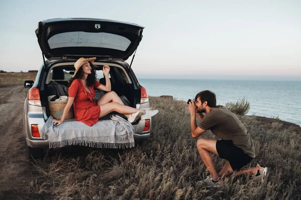 Чоловік фотографує жінку в червоному одязі і капелюсі, що сидить всередині відкритої частини автомобіля на задньому плані моря — стокове фото