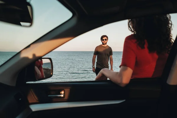 Vista a través de la ventana del coche, el hombre de pie frente a la mujer rizada en vestido rojo en el fondo del mar al atardecer —  Fotos de Stock