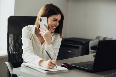 Ofiste çalışan kadın çalışan, akıllı telefondan bir telefon görüşmesi yaparken not alan kadın.