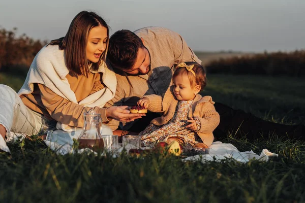 Młoda rodzina korzystająca z pikniku na świeżym powietrzu o zachodzie słońca, mama i tata z córeczką dziecka w weekend — Zdjęcie stockowe