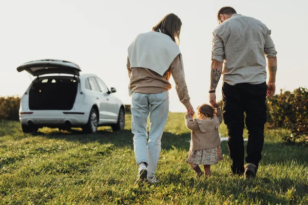 Молодая семья наслаждается поездкой на внедорожнике автомобиль, мать и татуированный отец помогая их ребенок дочь делает первые шаги на открытом воздухе в поле на закате — стоковое фото