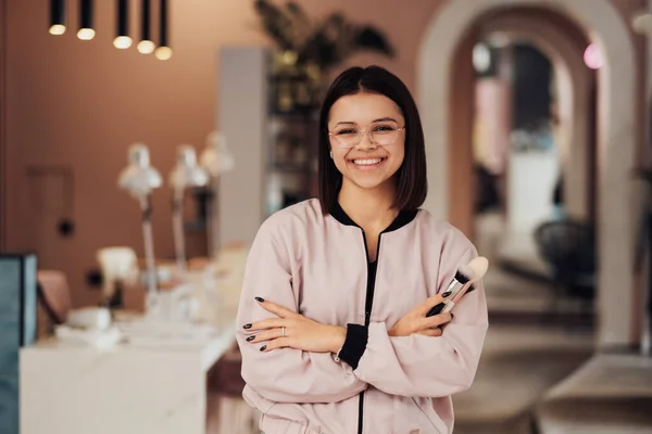 Retrato de joven alegre artista maquillaje femenino de pie en el salón de belleza, mujer caucásica en gafas con brazos doblados sosteniendo cepillos — Foto de Stock