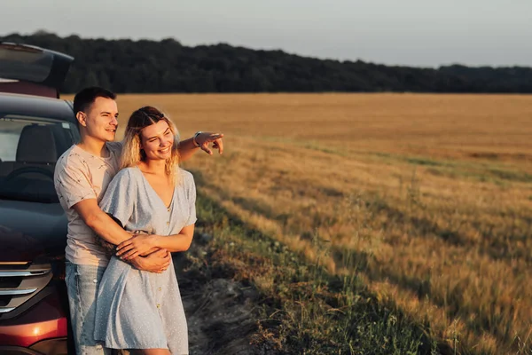 Wesoła młoda para przytulająca się w pobliżu samochodu, biała kobieta i mężczyzna cieszący się podróżą o zachodzie słońca, kopia miejsca — Zdjęcie stockowe