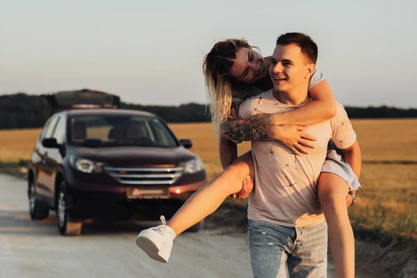 Caucasian Man Carries Girlfriend on His Back on the Background of Their SUV Car, Young Couple Enjoying Their Road Trip at Sunset — Stok Foto