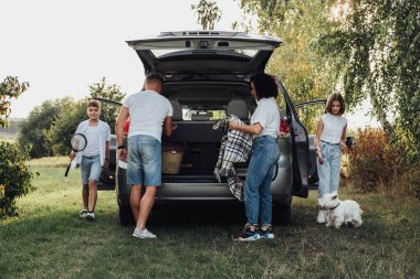 Minivan Car ile Dört Üye Aile Seyahati, Anne ve Baba ve İki Genç Çocuk ve Evcil Batı Highland Beyaz Teriyer Köpeği Açık Hava Pikniği Yapıyor
