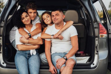 Mini arabanın bagajında oturan mutlu dört beyaz aile ferdinin portresi. Anne ve babası ve iki ergen çocuğu var. Oğlu ve kızı hafta sonu dışarı çıkıyor.