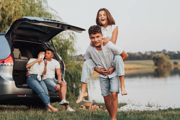 Двое счастливых детей-подростков играют на озере, в то время как их родители сидят в багажнике минивэна, четверо белых членов семьи наслаждаются поездкой на открытом воздухе