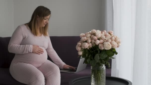 Размытая беременная женщина в декретном отпуске фрилансер Работает на ноутбуке на диване дома, букет цветов розы на переднем плане в фокусе — стоковое видео