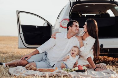 Bebekle birlikte gülümseyen genç aile şehrin dışında piknik yapıyor, anne ve baba kızlarıyla birlikte dışarıda oturan arabanın yanında oturan gibi giyiniyor.