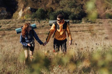 Gün batımında Tepede Yürürken Erkek ve Kadın El ele tutuşuyor Genç Çift Sırt çantaları ve Kamp Mat ile Yolculuklarının tadını çıkarıyor