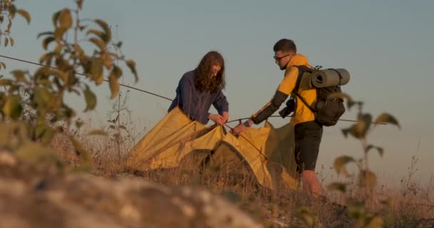 2人の若い旅行者が丘の上で夜を過ごすために日没時にテントを作る — ストック動画
