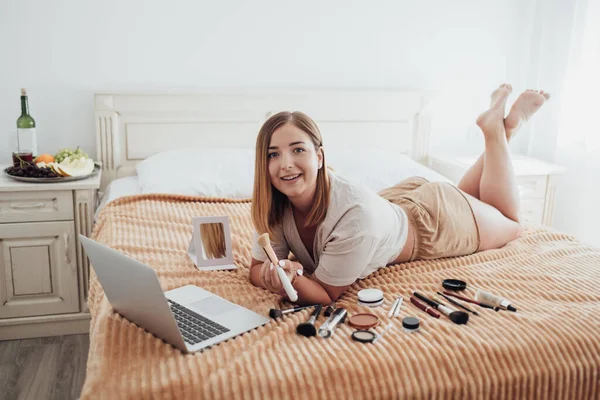 Young Caucasian Woman Laying on Bed with Cosmetics and Looking Into Camera During Her Online Master Class Through Laptop