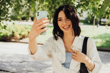 Neşeli Genç Kadın, Smartphone 'da Sunny Day' de parkta ağaçların arasında video çağrısı yapıyor.