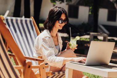 Güneş gözlüklü genç esmer kadın laptopta çalışıyor ve restoranın terasında otururken kokteyl içiyor, iş yerinde serbest çalışıyor.