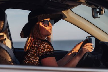 Gün batımında Şapkalı ve Güneş Gözlüklü Genç Neşeli Kadın Sürücü Aracı