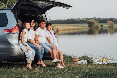 Minivan Car 'ın Bagajında oturan Kafkas Ailesi, Anne ve Baba ve İki Genç Çocuk, Oğul ve Kız Hafta Sonu Açık Hava Pikniği Yapıyor Göl kenarında