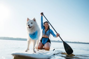 Cheerful Woman Paddleboarding with Her Pet on City Lake, Snow-White Japanese Spitz Dog Standing on Sup Board clipart