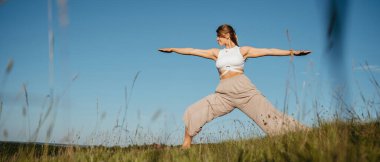 Young Woman Practicing Yoga Outdoors in the Field with Blue Sky on Background